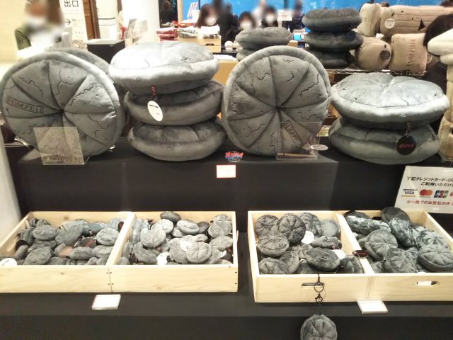 A display from a museum gift shop of plushies shaped like carbonized loaves of bread from Pompeii, i.e. round and segmented, with a stamps on them and cracks in them.