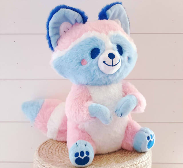 A plush raccoon, sitting up with paws curled inward. It's colored like the trans flag, with a soft pink body, white stomach, and eye/ear markings shades of light blue